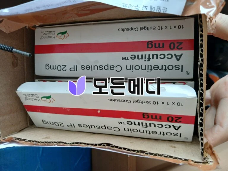 Inside a cardboard box, several white pharmaceutical boxes with a red band are stacked, showing small green-orange logo and upside-down text.