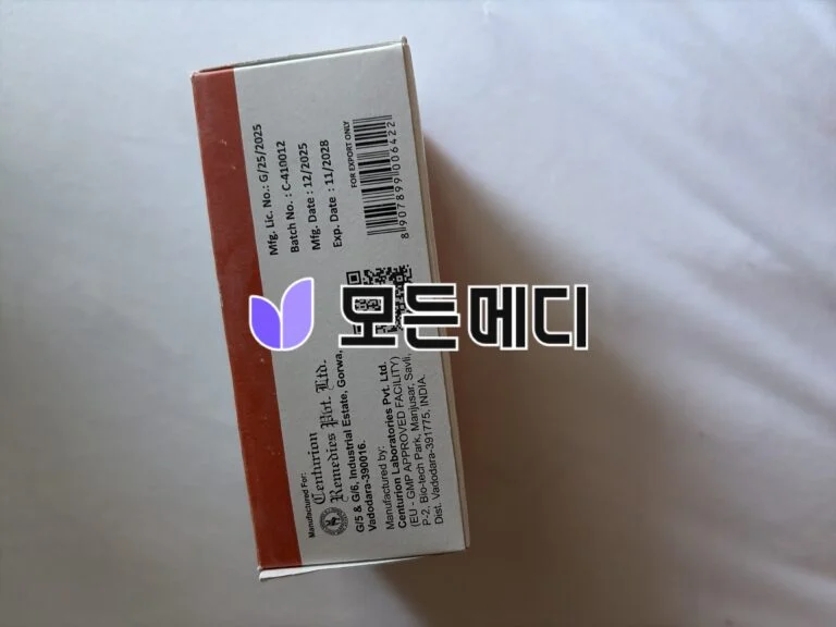 Tilted white and orange product box with a barcode and manufacturing details on the side, photographed against a light background.